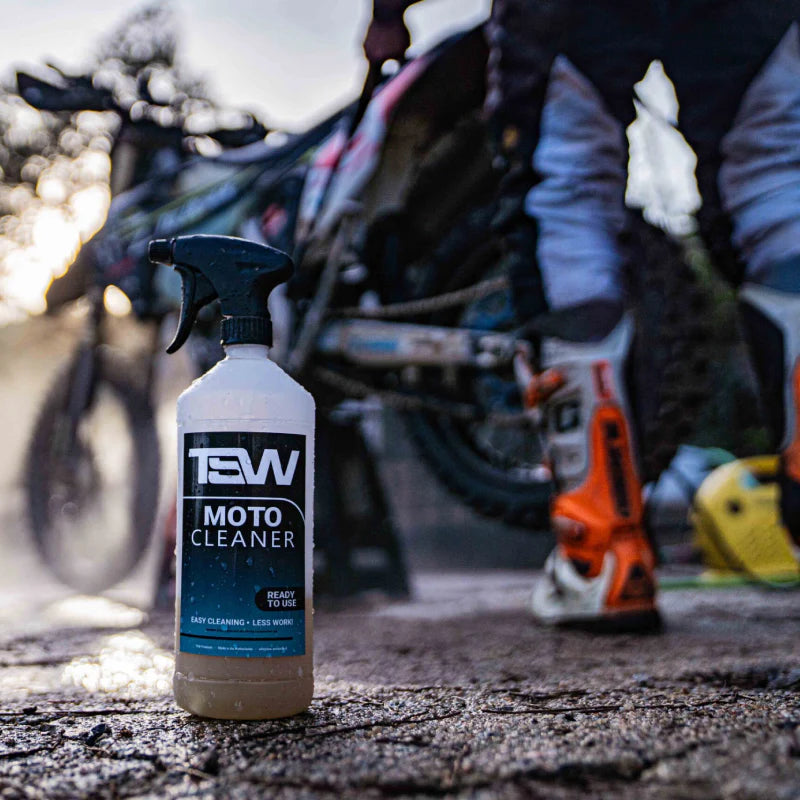Een fles TWS moto cleaner schoonmaak middel voor enduro en crossmotoren met op de achtergrond iemand die zijn motor met een hogedrukspuit schoonmaakt.